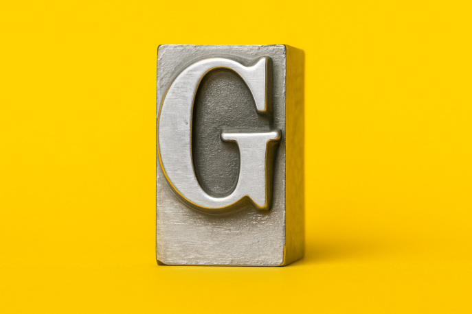 Nahaufnahme eines silbernen Metall-Letterpress-Buchstabenblocks mit dem Buchstaben G auf gelbem Hintergrund.