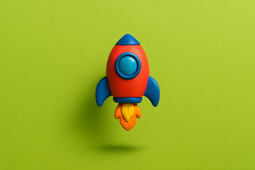 Eine kleine, bunte Spielzeugrakete schwebt zentral vor einem kräftigen grünen Hintergrund. Unter ihr liegt ein weicher Schatten, der den Eindruck erzeugt, dass sie kurz über dem Boden startet.