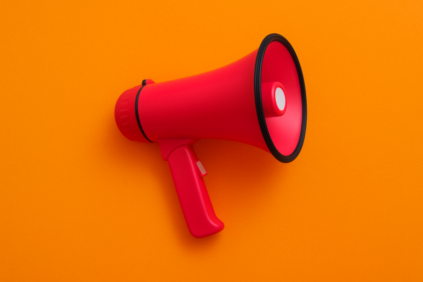 Ein pink-rot gefärbtes Megafon liegt seitlich auf einem kräftig orangefarbenen Hintergrund. Es wirkt modern und auffällig, als Symbol für eine wichtige Ankündigung oder neue Informationen.